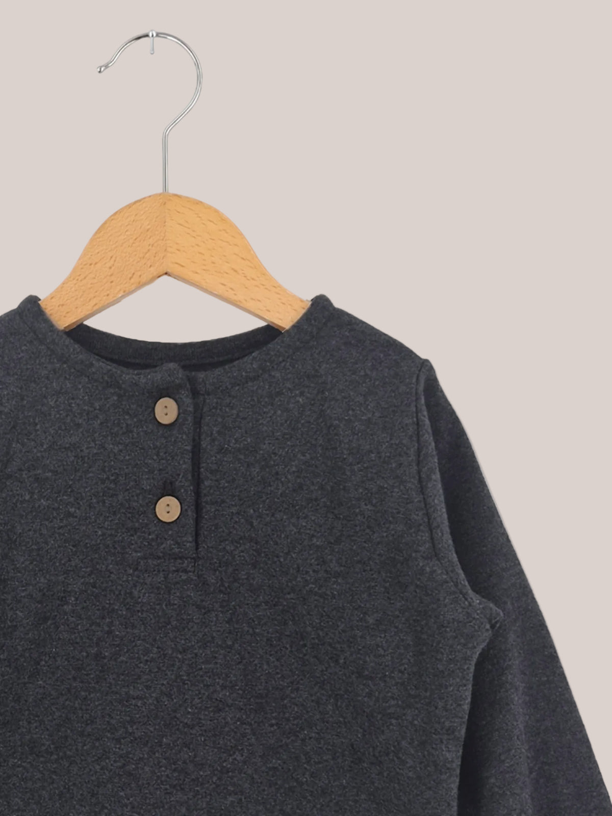 Canottiera in maglia fine - Fuliggine: un top a maniche lunghe color fuliggine con scollo rotondo e due bottoni di legno sull’abbottonatura, realizzato in morbido jersey di maglia fine, appeso a una semplice gruccia di legno.