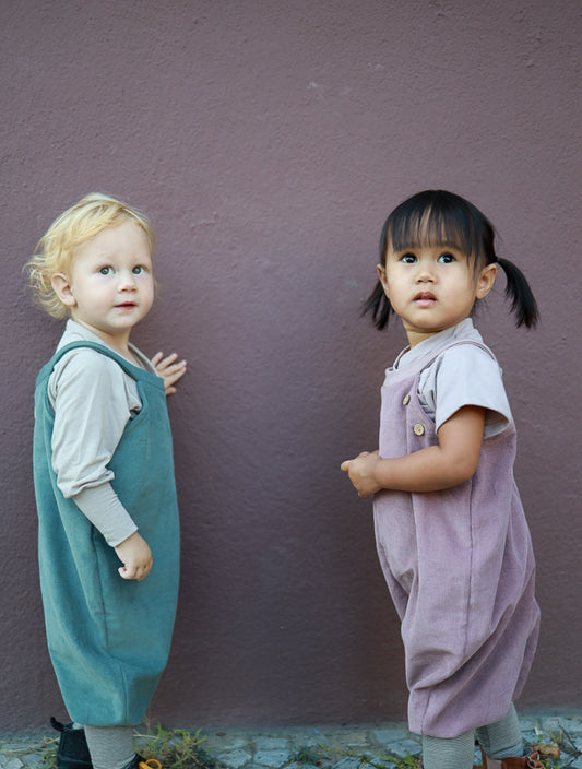 Two children stand next to each other; one is wearing the Feincord bib pants – Mauve, soft and comfortable corduroy dungarees without complicated fasteners, ideal for easy dressing and undressing. Sustainable children's clothing made in Berlin.