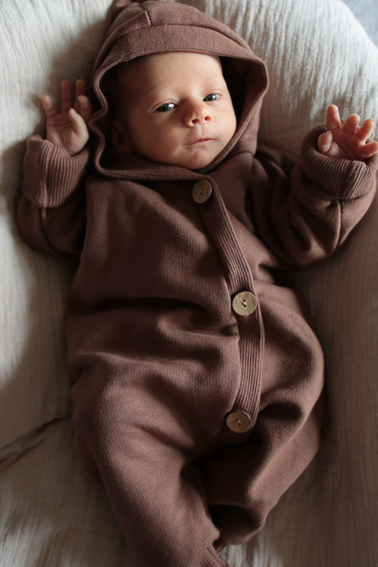 Ein lächelndes Baby liegt auf einem Bett, trägt gemütliche Baby- und Kleinkindkleidung, der Kuschelanzug Bio-Sweat , strampler baby von Petit Cochon aus Berlin.