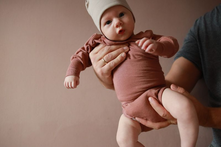 Eine Person hält ein Baby in einem zimtfarbenen Strampler. Der perfekte Baby body und die Erstausstattung für Babys.