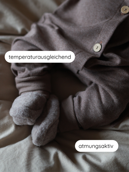 Babybooties Merino Wollfleece - Hafer: Ein Baby trägt einen braunen Einteiler und dicke, fleecige Booties aus atmungsaktivem, temperaturausgleichendem Merino-Wollfleece, ideal für warme, schweißfreie Füße.