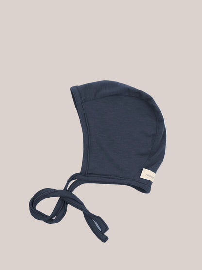 Baby Bonnet Cashmere Blend – Blueberry: A soft, navy-blue baby bonnet with long fabric ties, made from stretchy micromodal-cashmere fabric, ideal for gentle warmth and protection, with a small side label.
