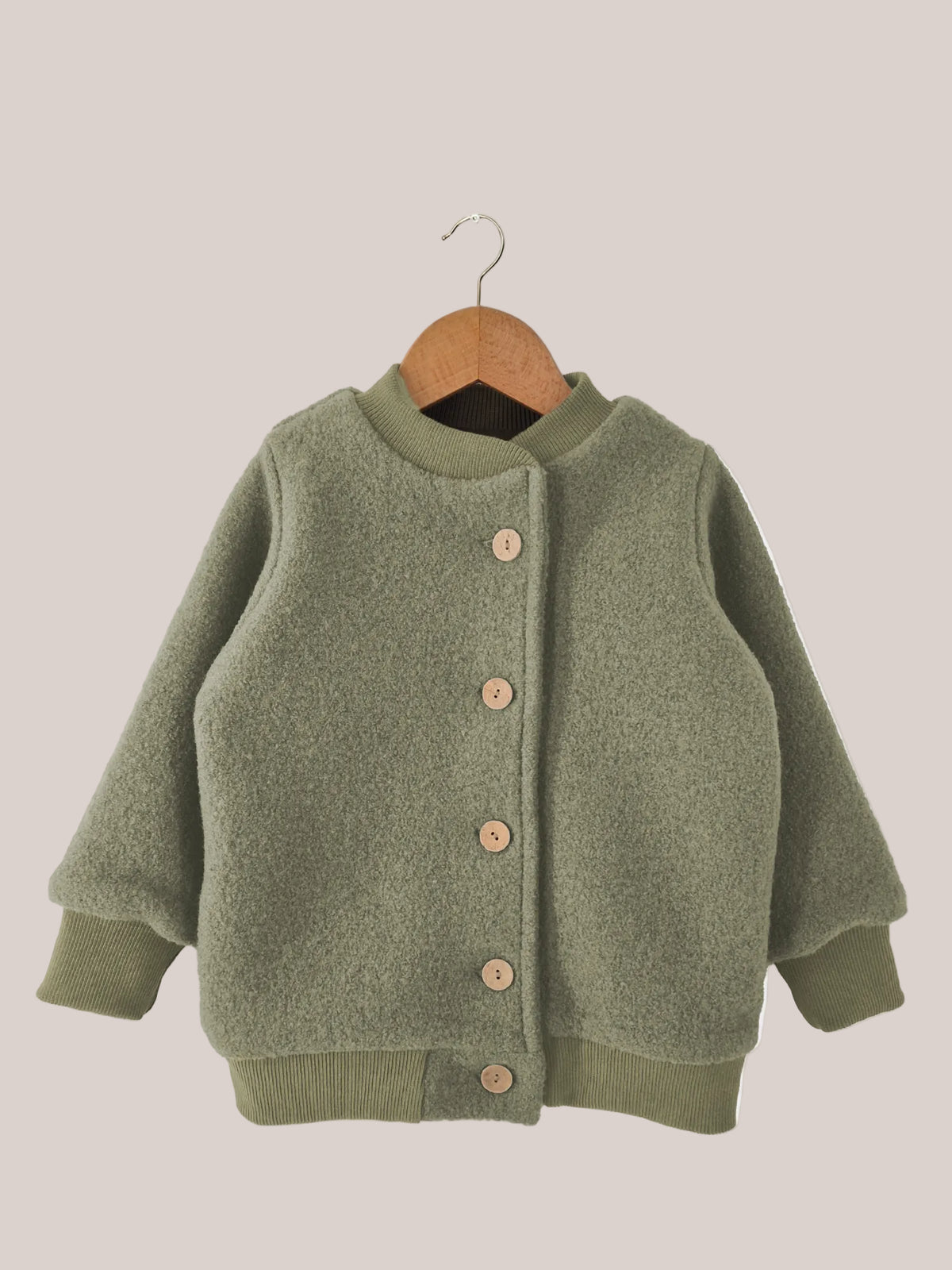 Veste bomber en laine foulée – pistache, présentée sur un cintre en bois, avec doublure en jersey et boutons, fabriquée de manière durable et conçue pour durer par PETIT COCHON.