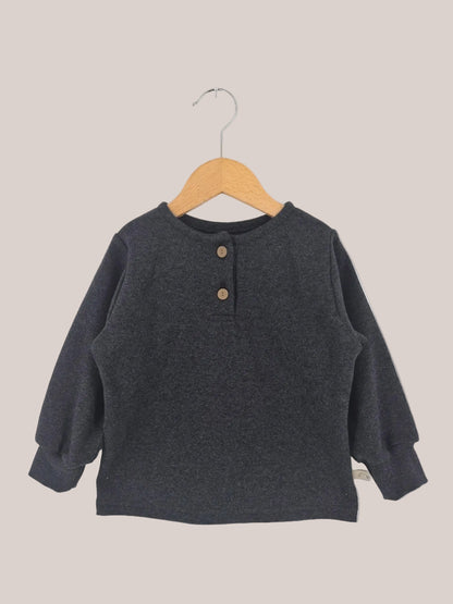 Maglietta in maglia fine - Fuliggine: una maglia Henley a maniche lunghe per bambini, in morbida maglia fine con scollo rotondo e due bottoni in legno, su una gruccia di legno davanti a uno sfondo bianco.