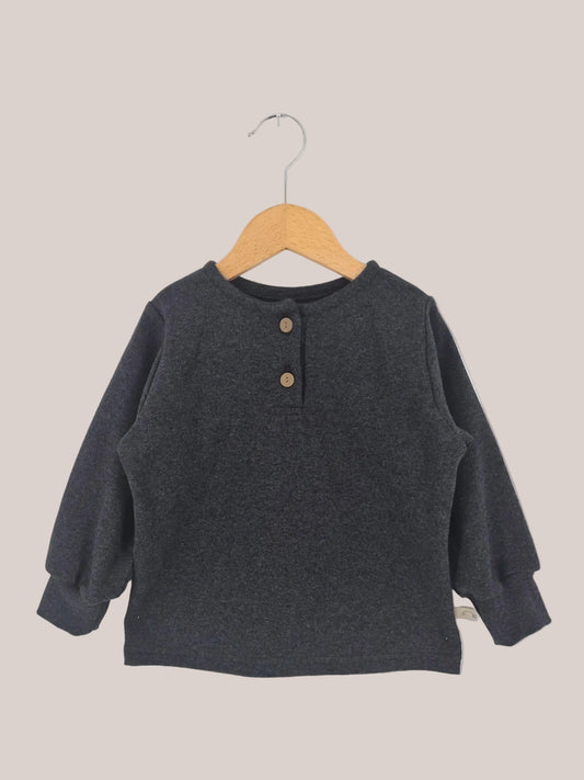 Hemdchen Feinstrick - Ruß: Ein langärmeliges Henley-Shirt für Kinder, aus weichem Feinstrick mit rundem Ausschnitt und zwei Holzknöpfen, auf einem Holzbügel vor weißem Hintergrund.
