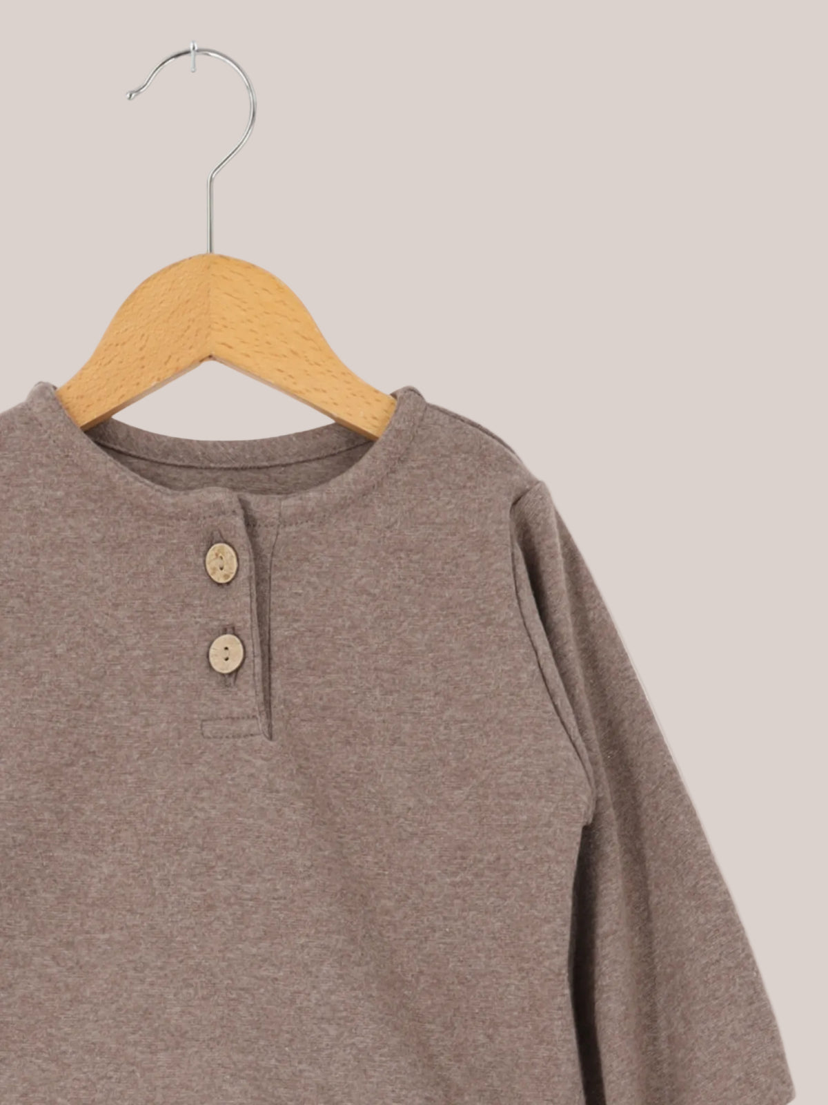 Maglietta in maglia fine - Praline: top a maniche lunghe per bambini con scollo rotondo e breve abbottonatura, realizzato in morbida maglia fine elasticizzata, appeso a una gruccia di legno su sfondo bianco.