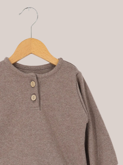 Maglietta in maglia fine - Praline: top a maniche lunghe per bambini con scollo rotondo e breve abbottonatura, realizzato in morbida maglia fine elasticizzata, appeso a una gruccia di legno su sfondo bianco.