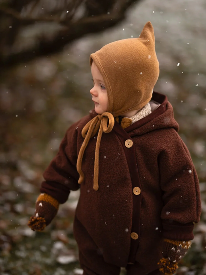 Tout-petit dans une combinaison douillette en laine bouillie – chocolat, vêtu d’une combinaison marron à boutons en bois, d’un bonnet en tricot jaune et de moufles assorties, debout sous une légère chute de neige. Parfait pour les saisons froides.