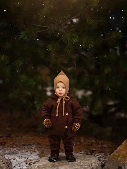 Tout-petit en combinaison douillette en laine foulée – chocolat, combinaison marron une pièce en laine avec boutons, moufles assorties et bonnet beige, debout sur une pierre dans une forêt hivernale, entouré de flocons de neige virevoltants.