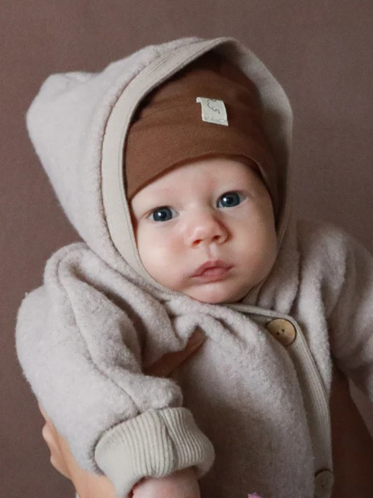 Kuschelanzug Wollwalk - Sand: Ein Baby im weichen, hellbeigen Wollwalkanzug mit großen Holzknöpfen und warmer Kapuze, getragen von Erwachsenen. Der Anzug schützt, wärmt und bietet Bewegungsfreiheit.