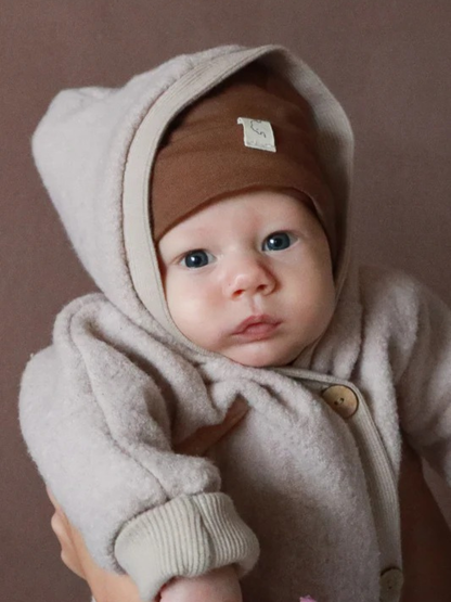 Kuschelanzug Wollwalk - Sand: Ein Baby im weichen, hellbeigen Wollwalkanzug mit großen Holzknöpfen und warmer Kapuze, getragen von Erwachsenen. Der Anzug schützt, wärmt und bietet Bewegungsfreiheit.