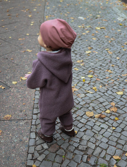 Kind trägt den Kuschelanzug Wollwalk - Malve aus Schurwolle, steht auf einem Ziegelweg. Der Overall hat eine Wichtelkapuze, enge Bündchen und bietet Bewegungsfreiheit und Schutz vor Wind. Wollwalk Anzug, Wollwalk Overall für Kinder, mitwachsender Wollwalk