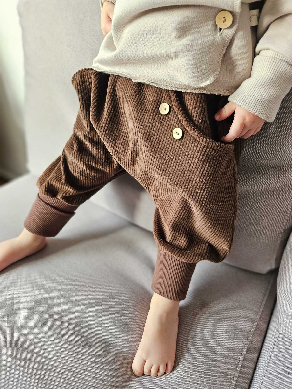 Kind in Knickerbocker Breitcord - Schoko, aus braunem Cord, mit Rippbündchen an den Knöcheln, Holzknöpfen an den Taschen, steht barfuß auf einem Sofa.