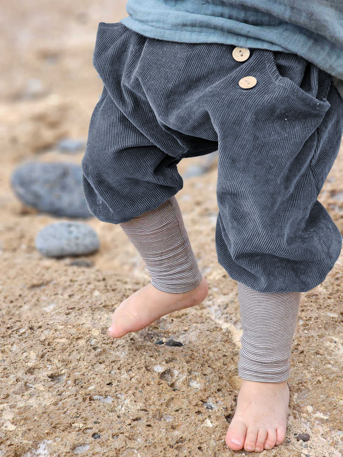 Un bambino è in piedi a piedi nudi sulla sabbia e indossa i pantaloni alla zuava in velluto a coste fine a righe color mirtillo; sono visibili i pantaloni di velluto dalla vestibilità comoda, con bottoni in legno, e i leggings a righe sotto, ideali per piccoli esploratori attivi.