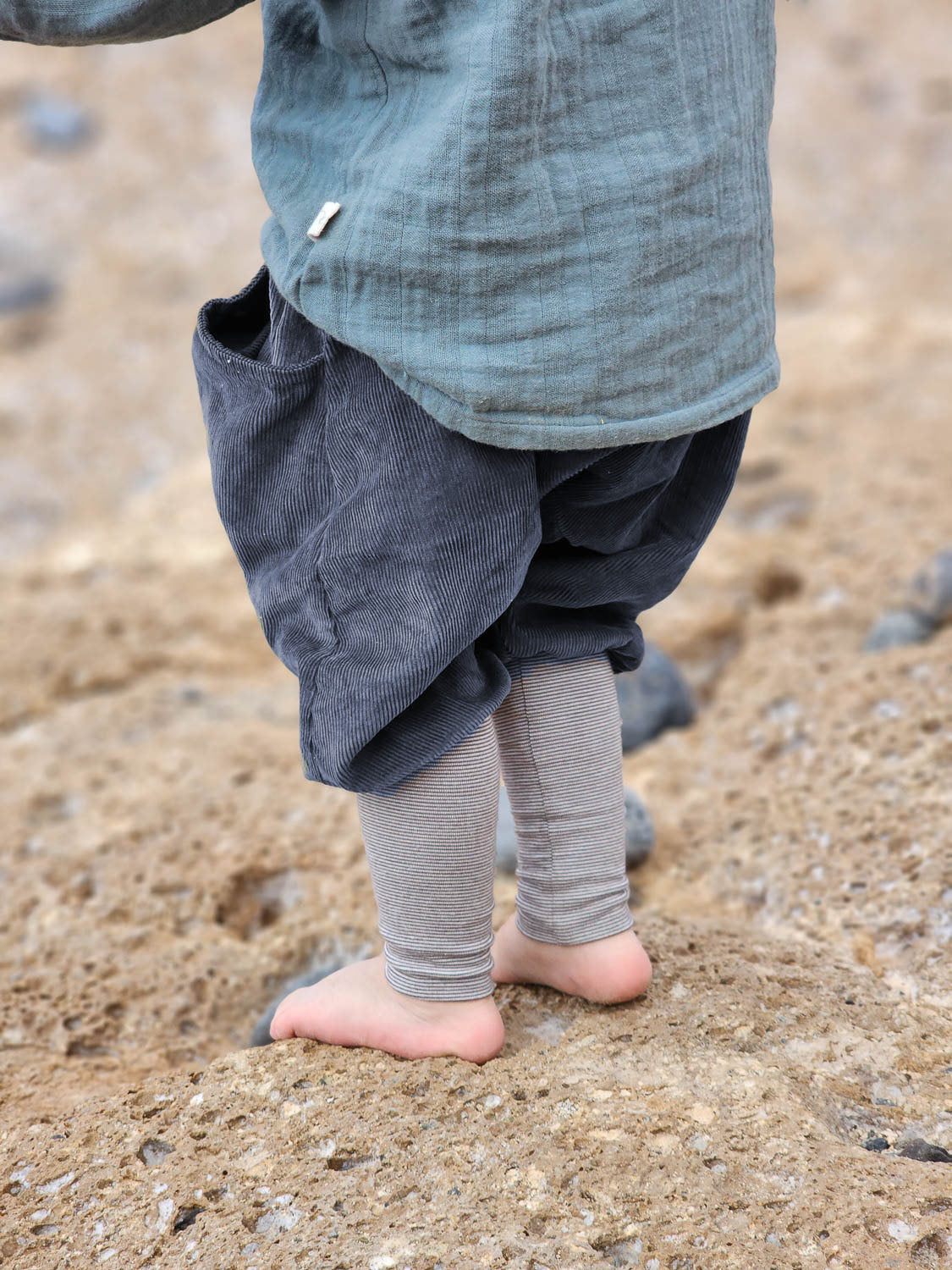 Knickerbocker Feincord Ringel - Mirtillo: primo piano di un bambino in piedi a piedi nudi su un terreno sabbioso, che indossa morbidi pantaloni di velluto a coste grigio scuro con fascia a righe, ideali per muoversi comodamente.