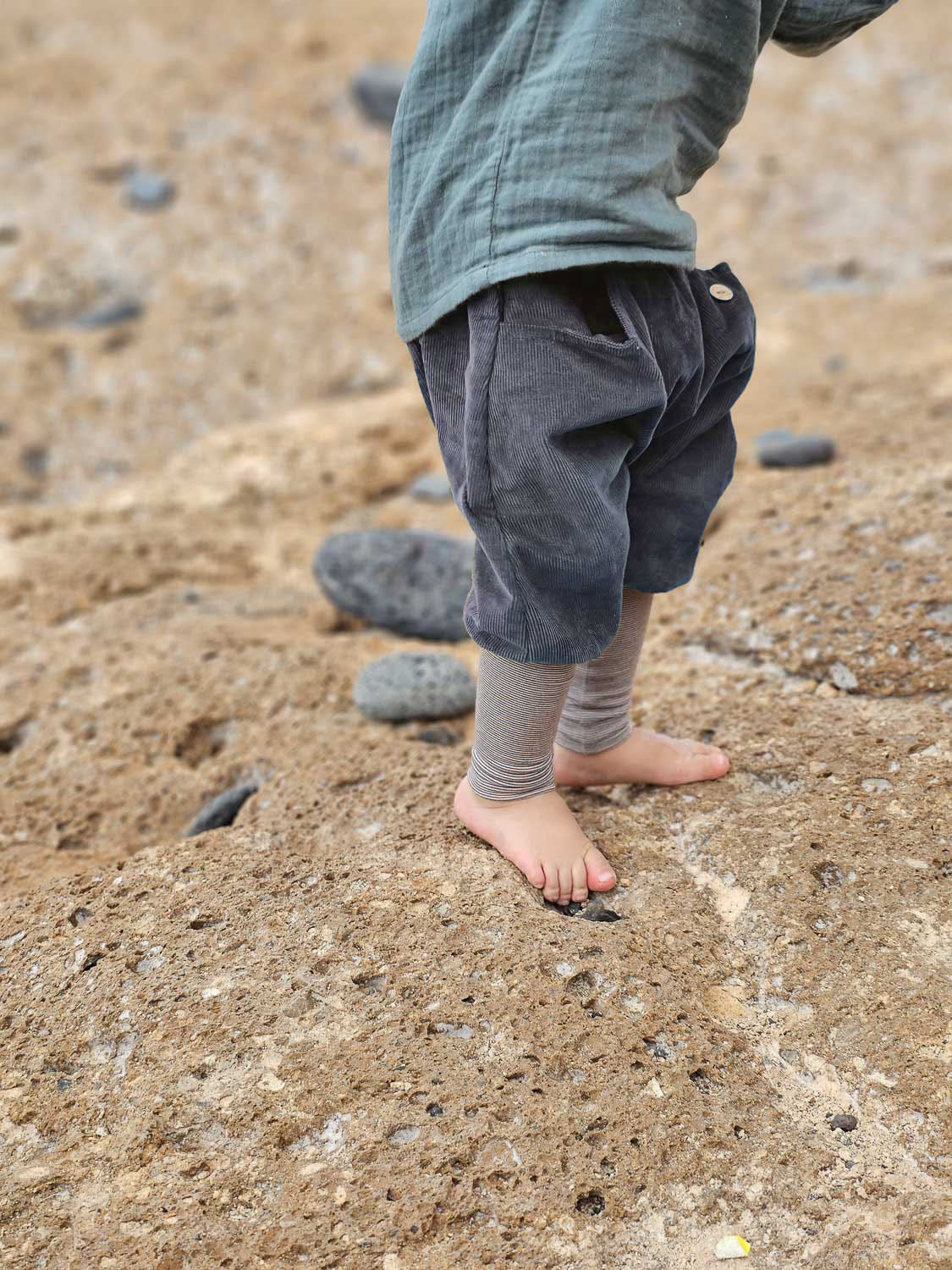 Bambino piccolo indossa i pantaloncini alla zuava in velluto a coste fini a righe – mirtillo, pantaloncini grigi in velluto a coste con fascia a righe, in piedi su un terreno sabbioso, che dimostrano libertà di movimento e comfort per piccoli esploratori e piccole esploratrici.