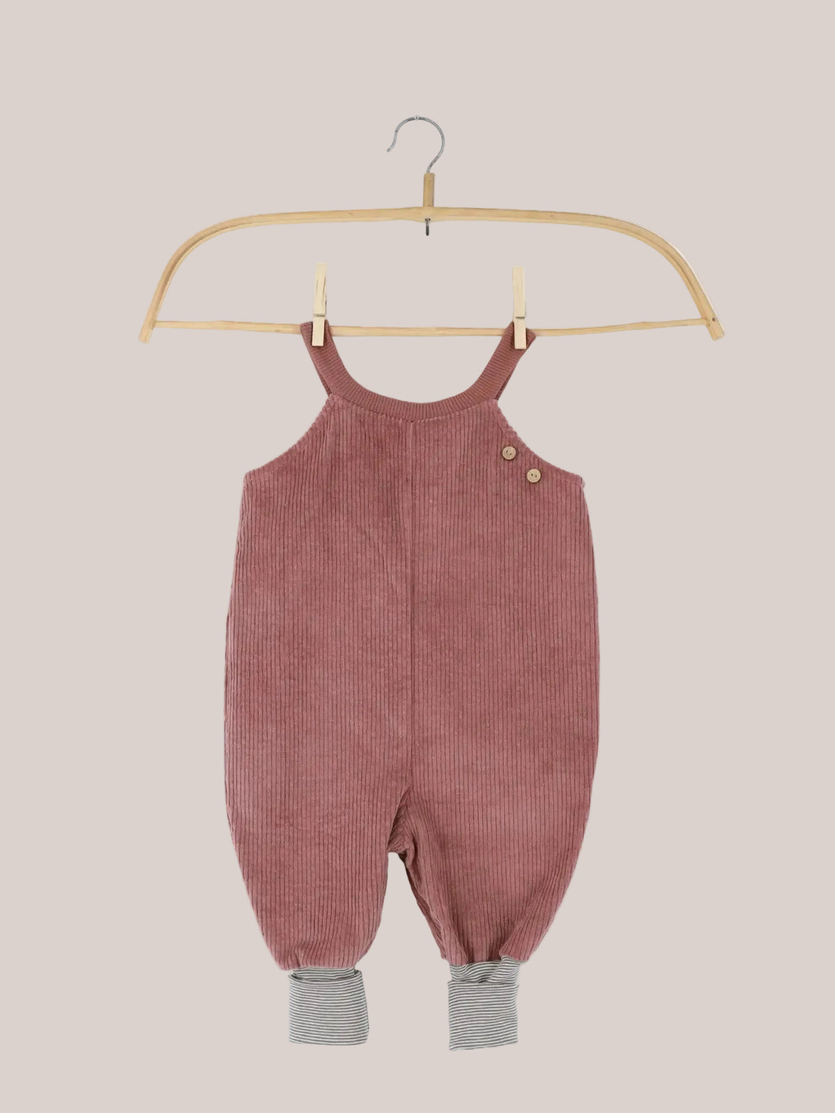 Peto infantil de pana ancha con espacio para el pañal – rosa palo: un peto tipo pelele de pana rosa empolvado para bebés y niños pequeños, con botones de madera en los tirantes y puños de rayas gris y blanco en las perneras, colgado en una percha de madera.
