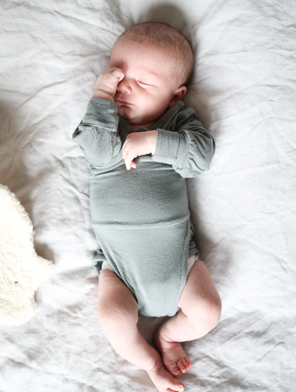 Neugeborenes trägt Loopbody Kaschmir-Mix - Taube, liegt auf weißem Laken. Der Einteiler hat lange Ärmel, ist knopf- und nahtlos, bietet sanften Komfort und passt sich dem Baby perfekt an.