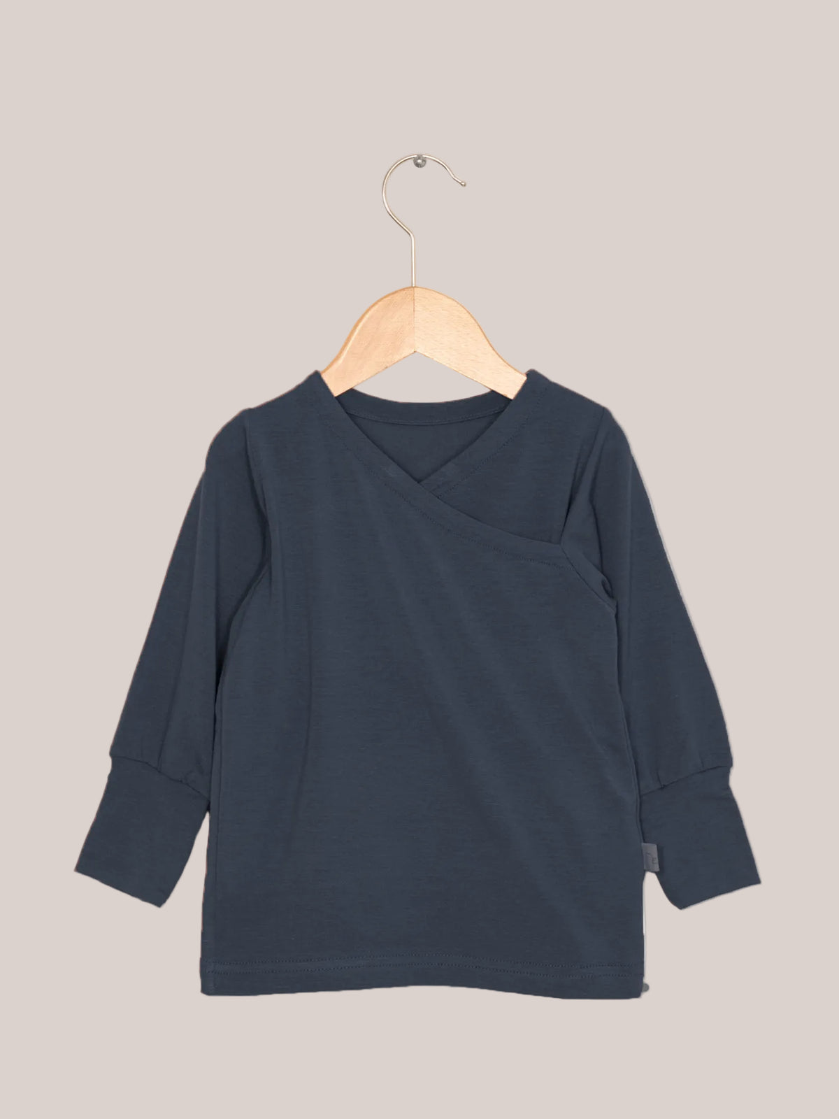 Wickelshirt aus Kaschmir-Mix - Blaubeere: Ein langärmeliges, dunkelblaues Shirt mit kimonoartigem Ausschnitt und gerippten Bündchen, hängend auf einem Holzkleiderbügel vor hellem Hintergrund.