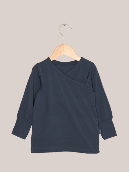Wickelshirt aus Kaschmir-Mix - Blaubeere: Ein langärmeliges, dunkelblaues Shirt mit kimonoartigem Ausschnitt und gerippten Bündchen, hängend auf einem Holzkleiderbügel vor hellem Hintergrund.