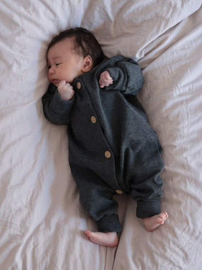 Baby in einem petit cochon Mini Overall aus dunkelgrauem, meliertem Feinstrick-Jersey mit Holzknöpfen auf dem Bett.
