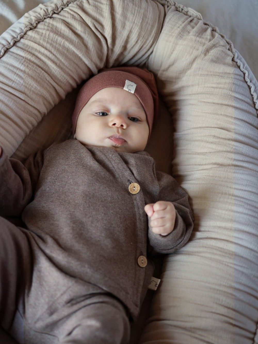 Baby in braunem petit cochon Wickeloberteil aus Feinstrick mit Kokosknöpfen, liegend in einem Babynest.