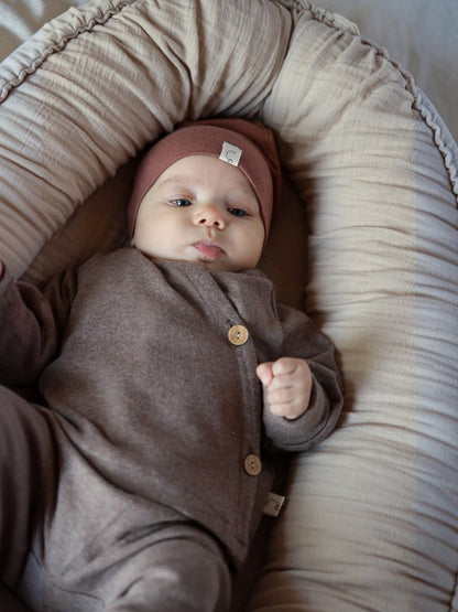 Baby in braunem petit cochon Wickeloberteil aus Feinstrick mit Kokosknöpfen, liegend in einem Babynest.