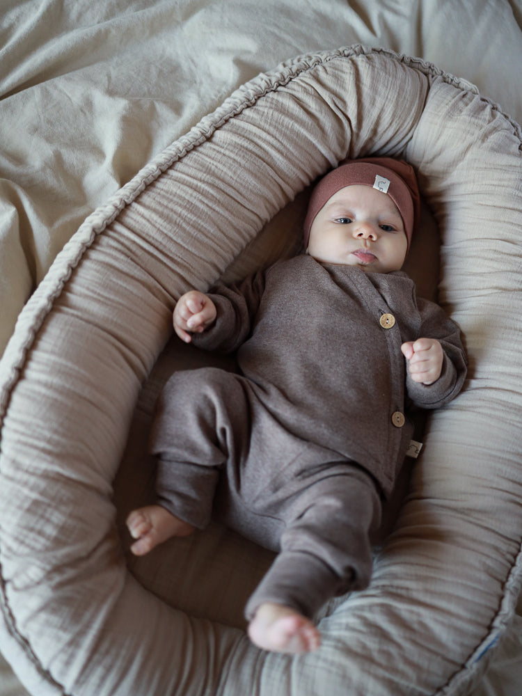 Baby in braunem petit cochon Schlüttli Wickeloberteil aus Feinstrick mit Kokosknöpfen in einem Babynest