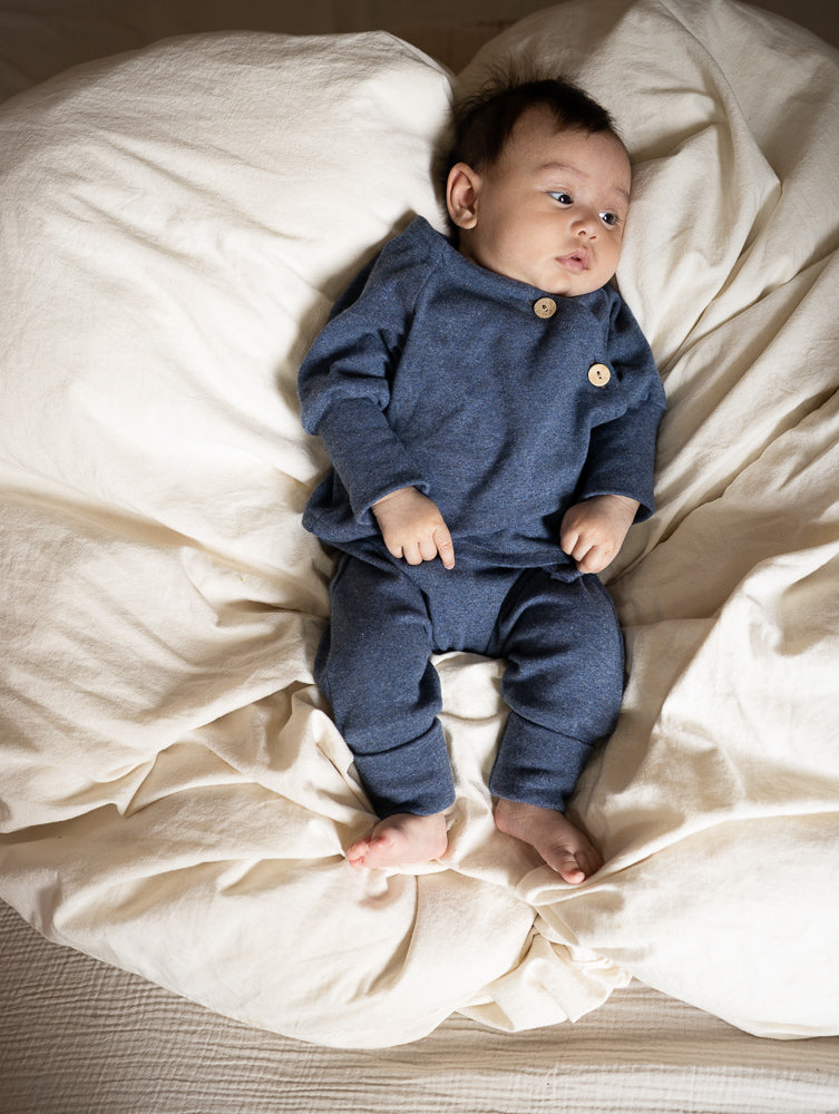 Baby von petit cochon in blauem Feinstrick-Schlüttli mit Kokosknöpfen, liegend auf einer hellen Decke.