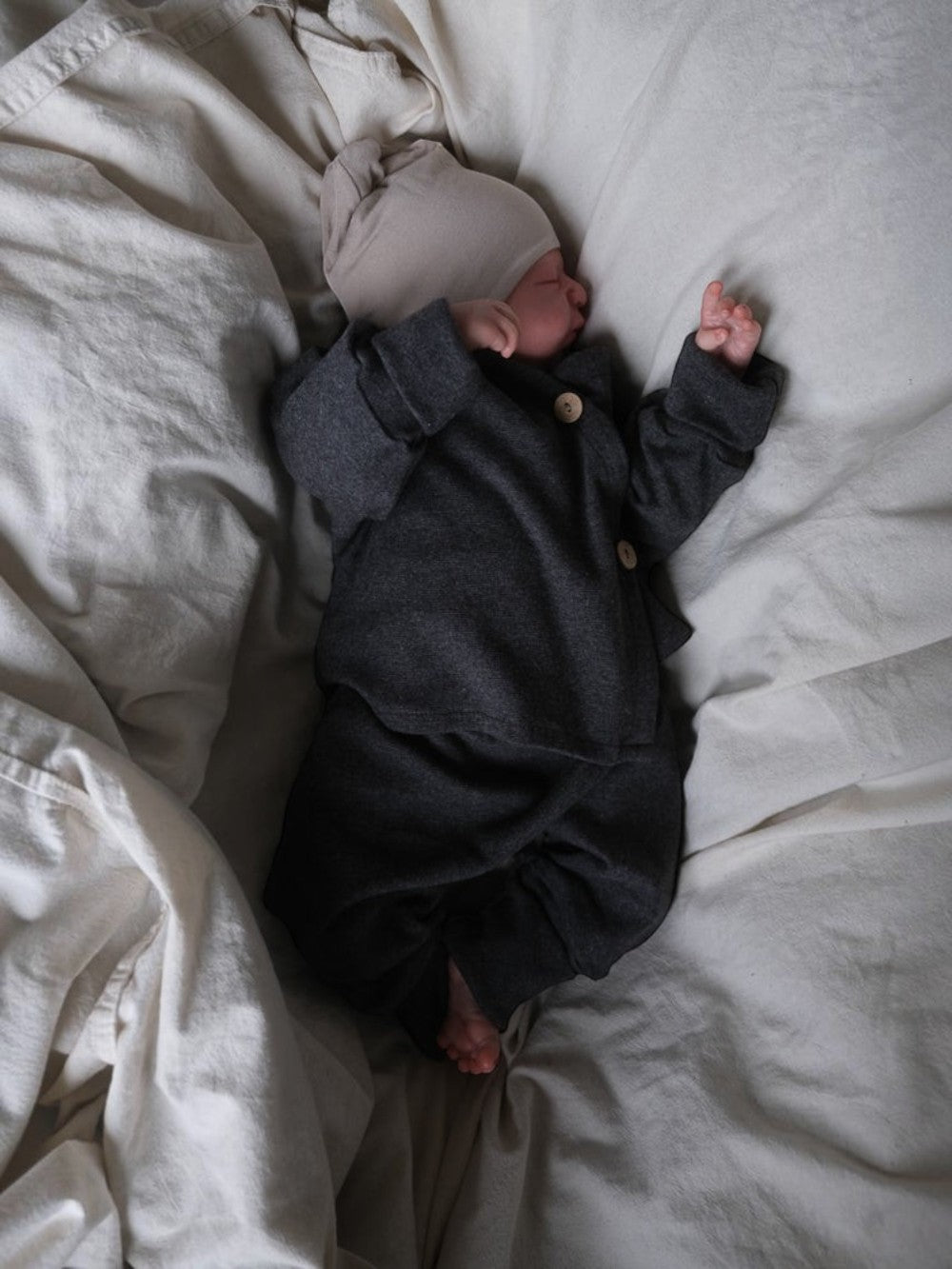 Neugeborenes in grauem petit cochon Baby Schlüttli aus weichem Feinstrick mit Kokosknöpfen auf heller Decke