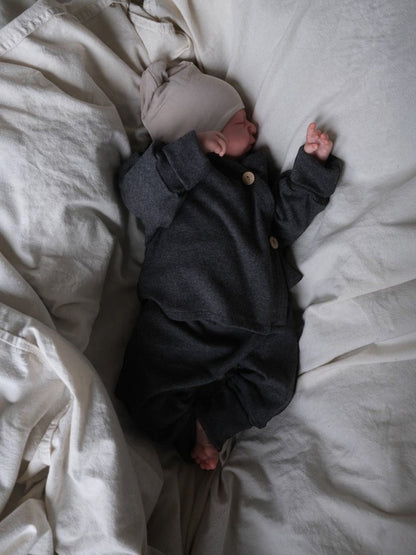 Neugeborenes in grauem petit cochon Baby Schlüttli aus weichem Feinstrick mit Kokosknöpfen auf heller Decke
