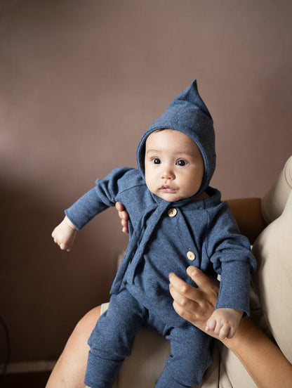 Baby von petit cochon in blauem Feinstrick-Schlüttli mit passender Zipfelmütze und Kokosknöpfen auf dem Arm gehalten