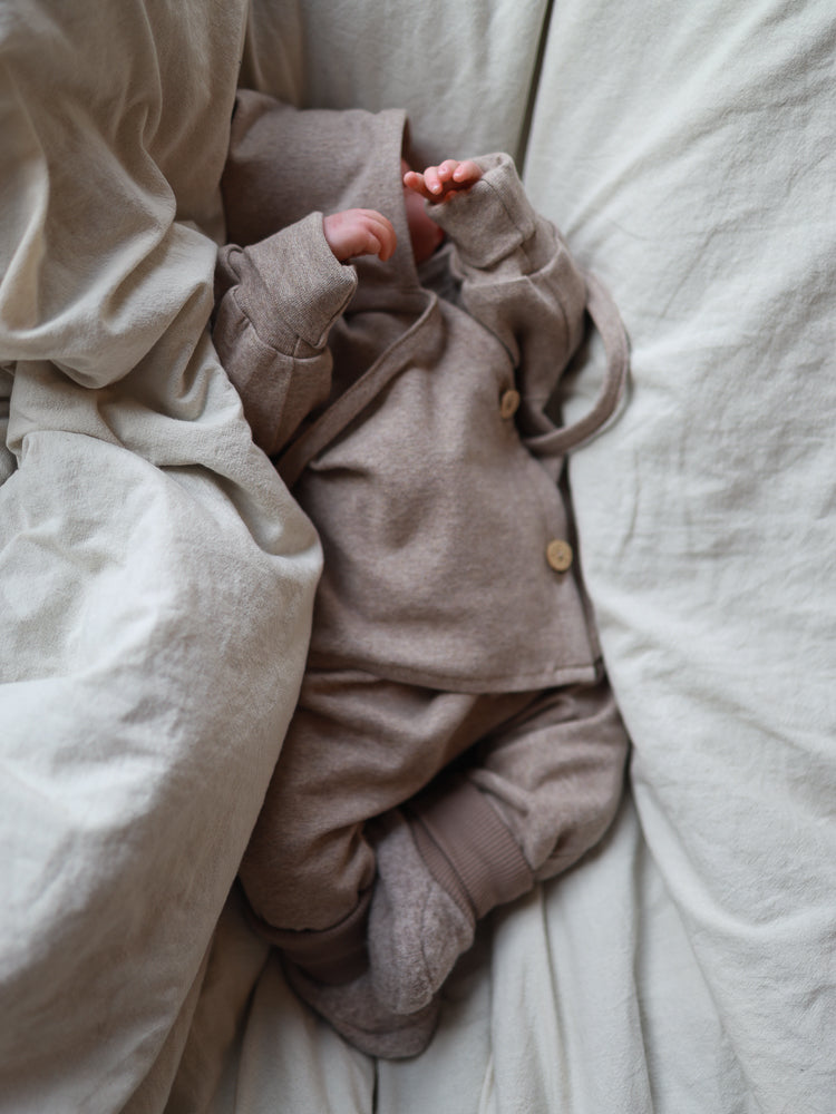 petit cochon Babyknicker aus weichem, meliertem Feinstrick-Jersey in Beige, getragen von einem Baby im Bett