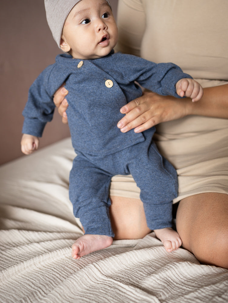 petit cochon Babyknicker aus blauem, meliertem Feinstrick-Jersey mit Holzknöpfen, getragen von einem Baby.