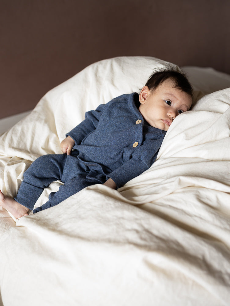 Baby in petit cochon Babyknicker aus blauem, meliertem Feinstrick-Jersey mit Holzknöpfen auf weicher Decke liegend