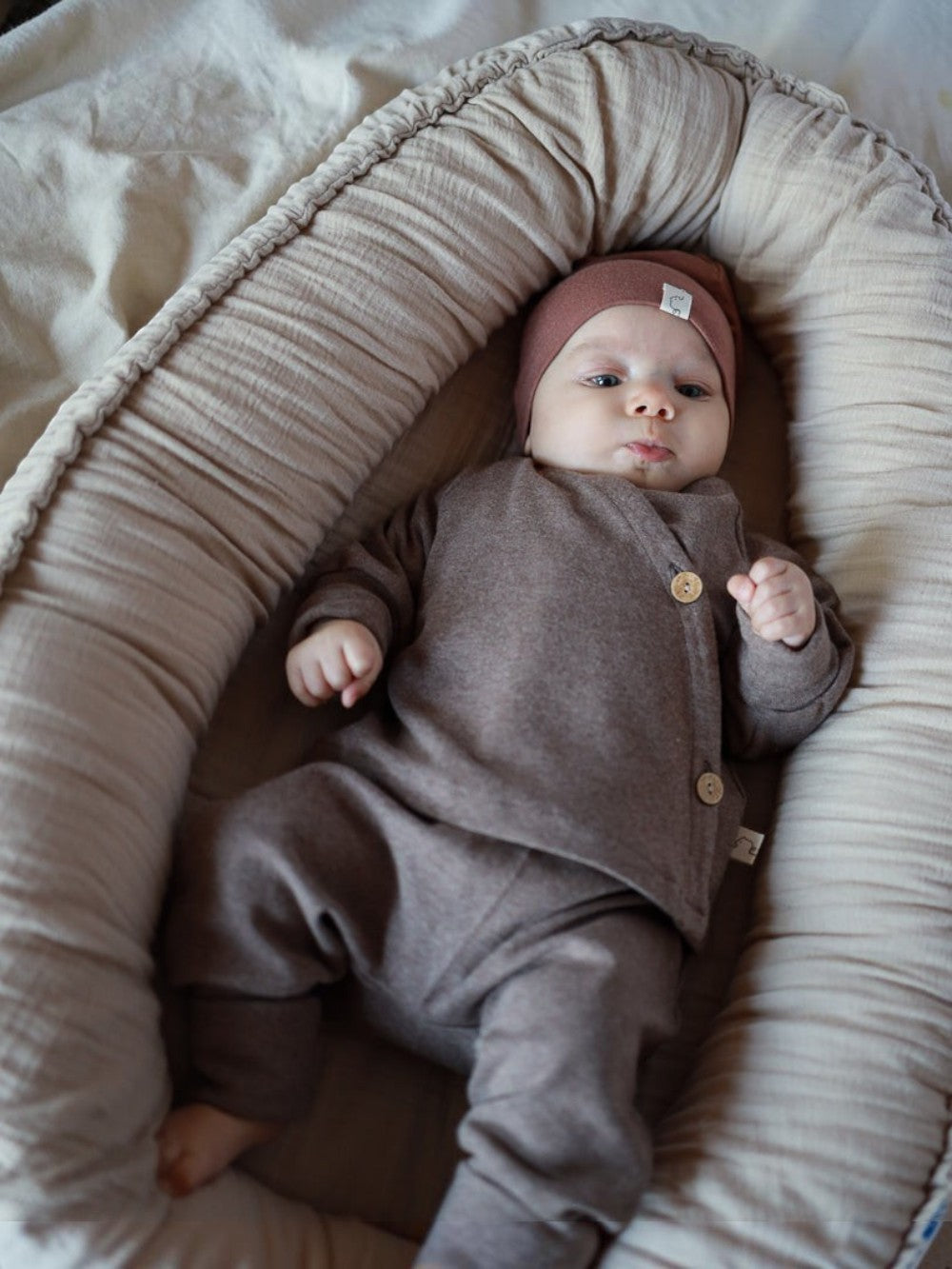 Baby in petit cochon Feinstrick-Babyknicker und Mütze, liegend in einem beigen Babynest aus weichem Jersey