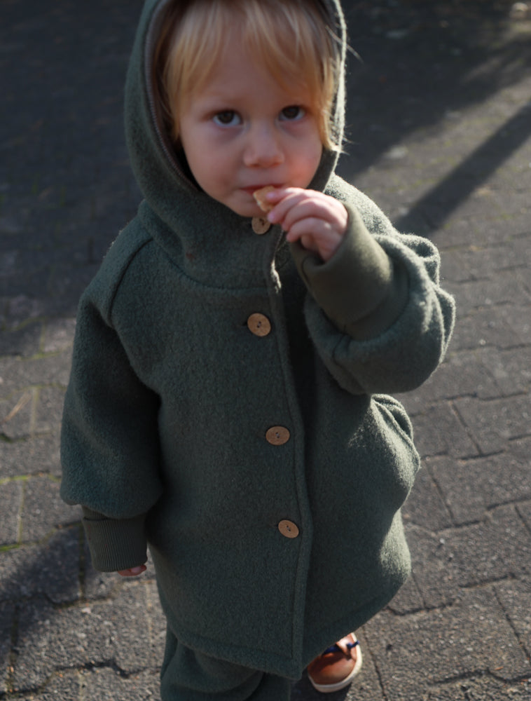 Kind trägt eine olivgrüne petit cochon Wichteljacke aus Wollwalk mit Kapuze und Holzknöpfen im Freien.