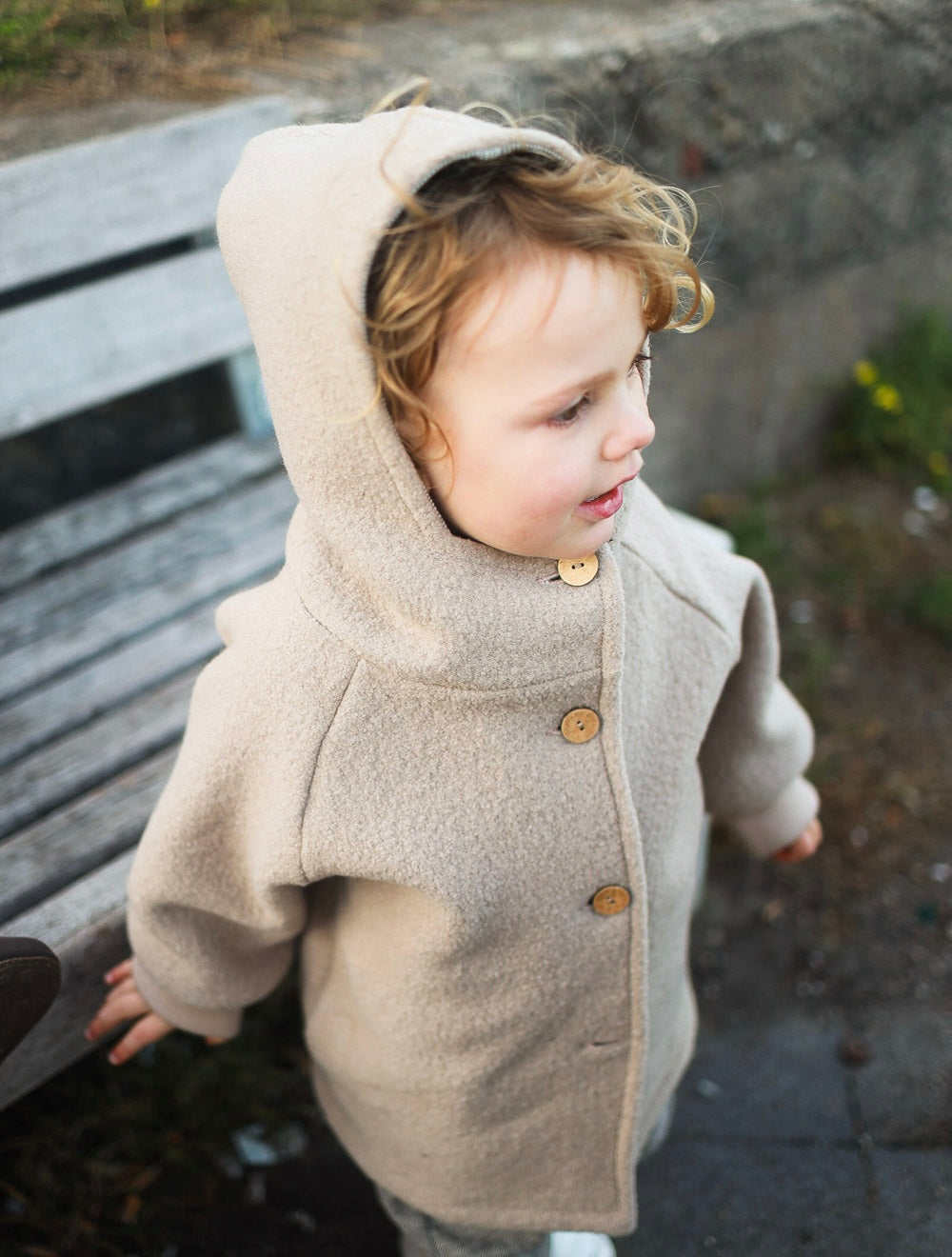 Kind trägt eine beige petit cochon Wichteljacke aus Wollwalk mit Kapuze und Holzknöpfen im Freien.