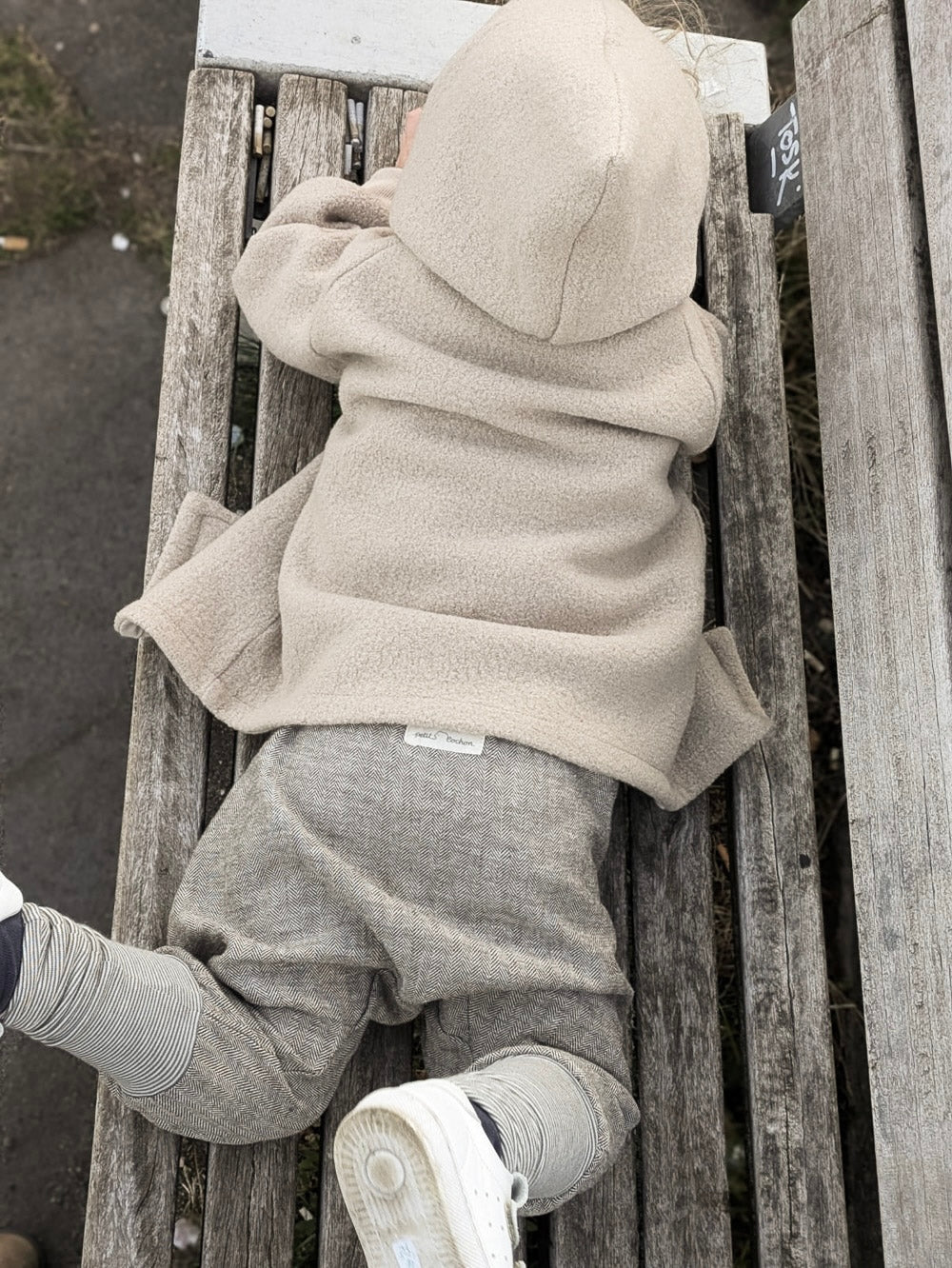 petit cochon Wichteljacke aus beigem Wollwalk mit Kapuze, getragen von einem Kind auf einer Holzbank im Freien