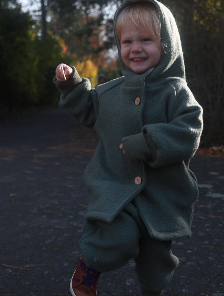 Ein Kind trägt eine grüne petit cochon Wichteljacke aus Wollwalk mit Kapuze und krempelbaren Bündchen im Freien.