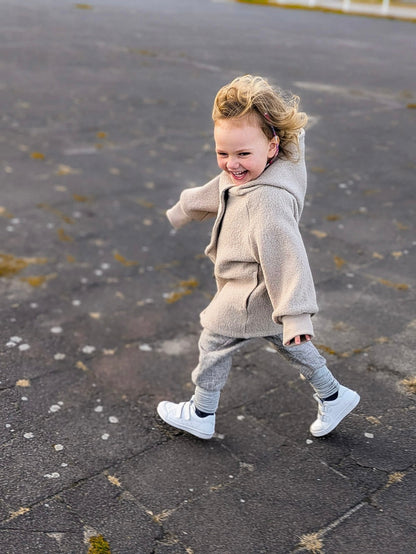 Ein Kind trägt eine beige petit cochon Wollwalk-Wichteljacke mit Kapuze und krempelbaren Bündchen beim Spielen im Freien.