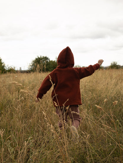 Kind in einer dunkelroten petit cochon Wollwalk-Wichteljacke mit Kapuze beim Spielen auf einer herbstlichen Wiese