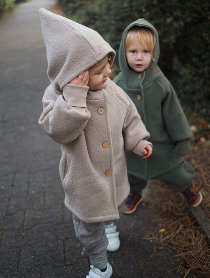 Zwei Kinder tragen petit cochon Wichteljacken aus beigem und olivgrünem Wollwalk mit Kapuze und Holzknöpfen im Freien.