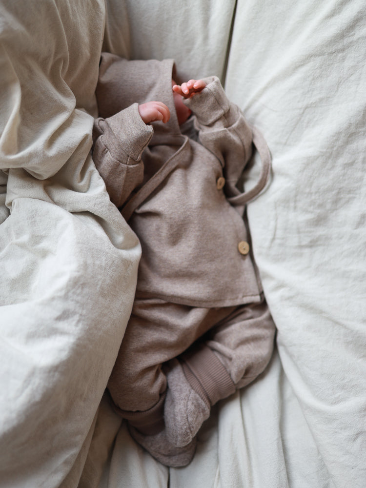 Neonato su un tessuto morbido, indossa il completo per bebè in maglia fine – avena mélange: giacca con cappuccio e bottoni in legno, pantaloni coordinati e scaldapiedi. Mani e gambe visibili in un ambiente naturale e rilassante.