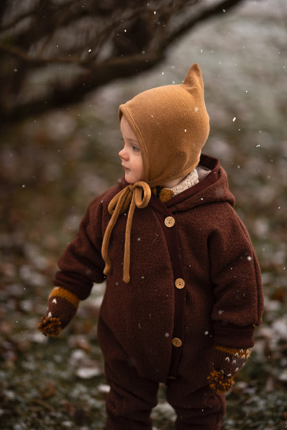 Enfant en combinaison douillette en laine foulée – châtaigne avec bonnet de lutin en tricot, idéale pour les saisons fraîches. La combinaison régule la chaleur et offre une grande liberté de mouvement, parfaite pour les activités en plein air.