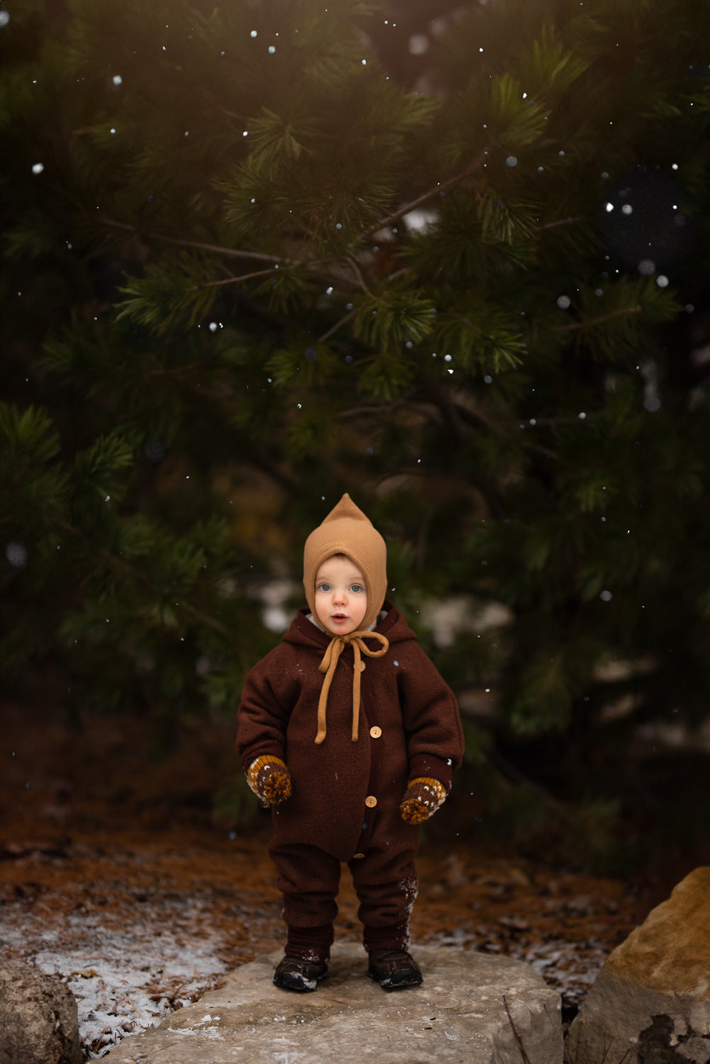 Un enfant porte la combinaison douillette en laine bouillie – châtaigne, en 100 % laine vierge, dans la neige, idéale pour les saisons fraîches grâce à ses propriétés chaudes et respirantes.