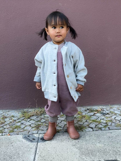 Mädchen trägt Schlupflatzhose Feincord - Malve vor lila Wand. Bequeme, handgenähte Latzhose aus weichem Cord, ideal für Kinder in verschiedenen Altersstufen.